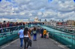 A busy pedestrian bridge with people walking towards the camera, under a cloudy sky with city buildings in the background.