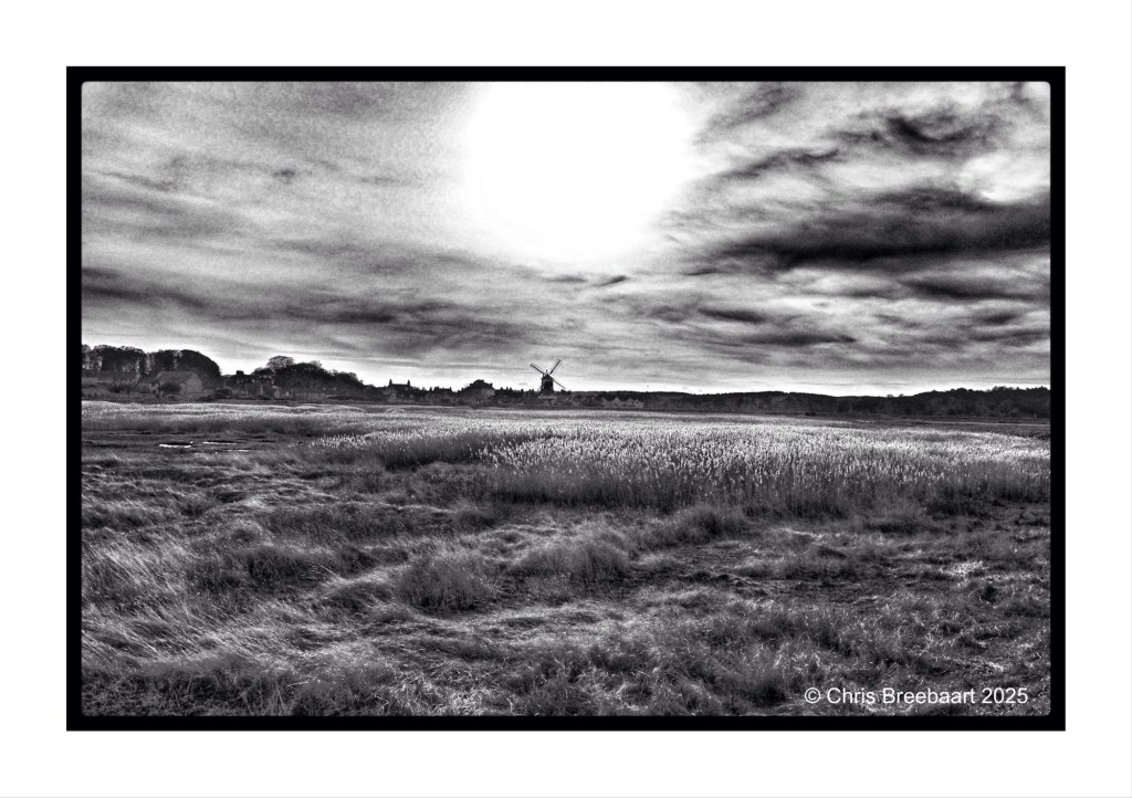 A black and white landscape image featuring Cley windmill in the distance, surrounded by grassy fields and a cloudy sky.