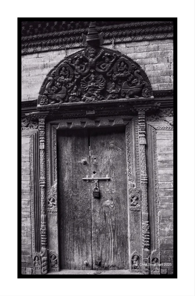 A detailed black and white photograph of a wooden door adorned with intricate carvings and a padlock, showcasing traditional architectural elements.