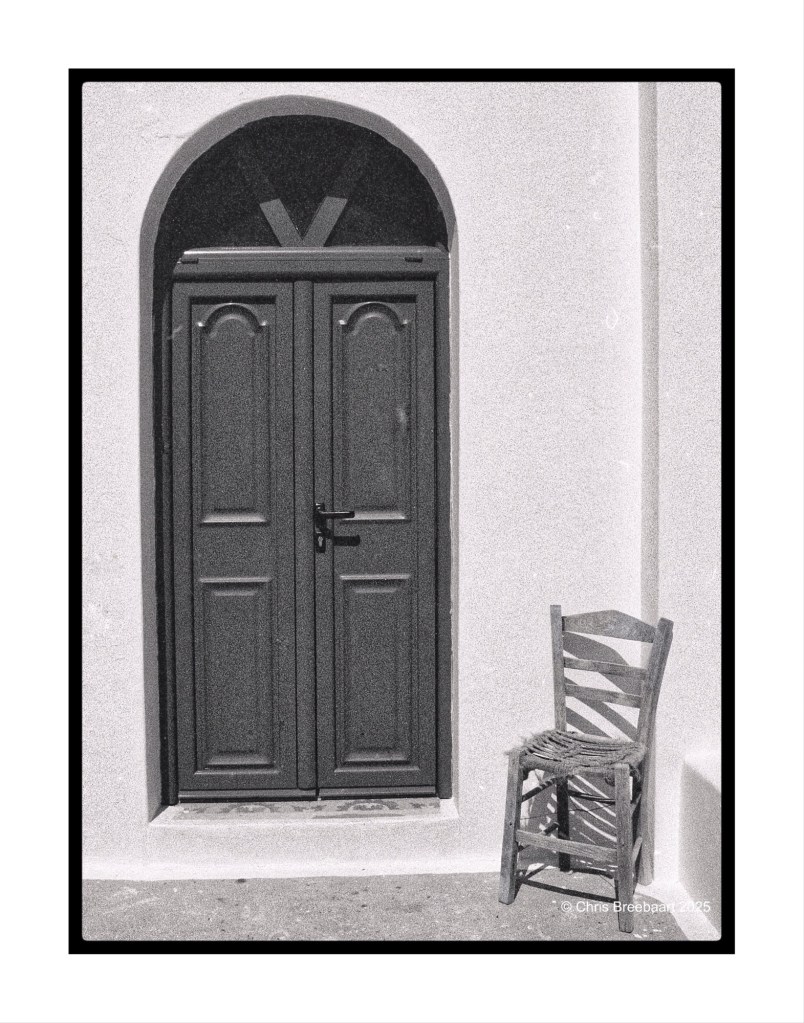 A black and white photograph of a double door with a simple design, partially open, situated beside a wooden chair against a white wall.