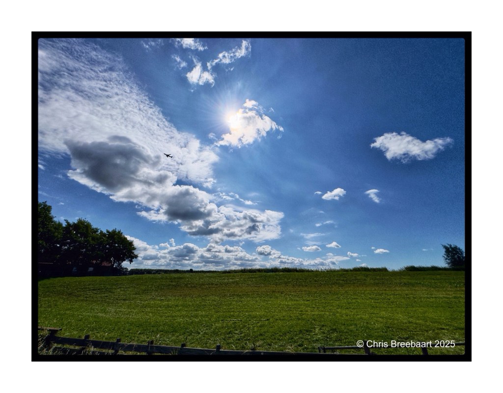 A wide blue sky filled with white clouds, with sunlight shining down over a lush green field.