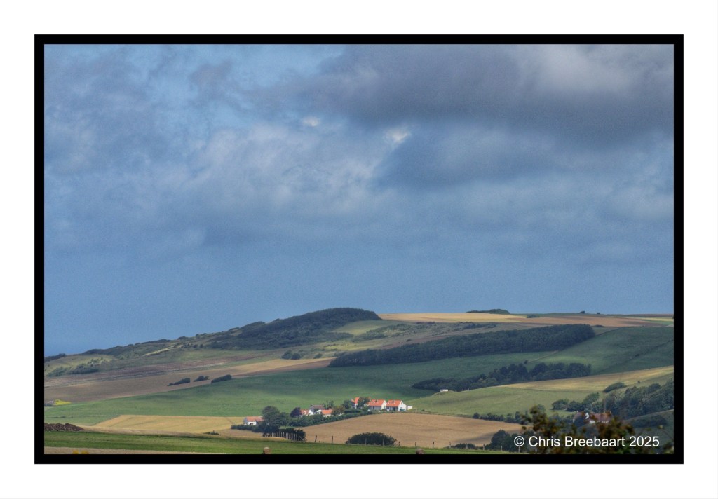 A panoramic view of rolling green and yellow fields under a cloudy sky, with a small collection of houses visible in the distance.
