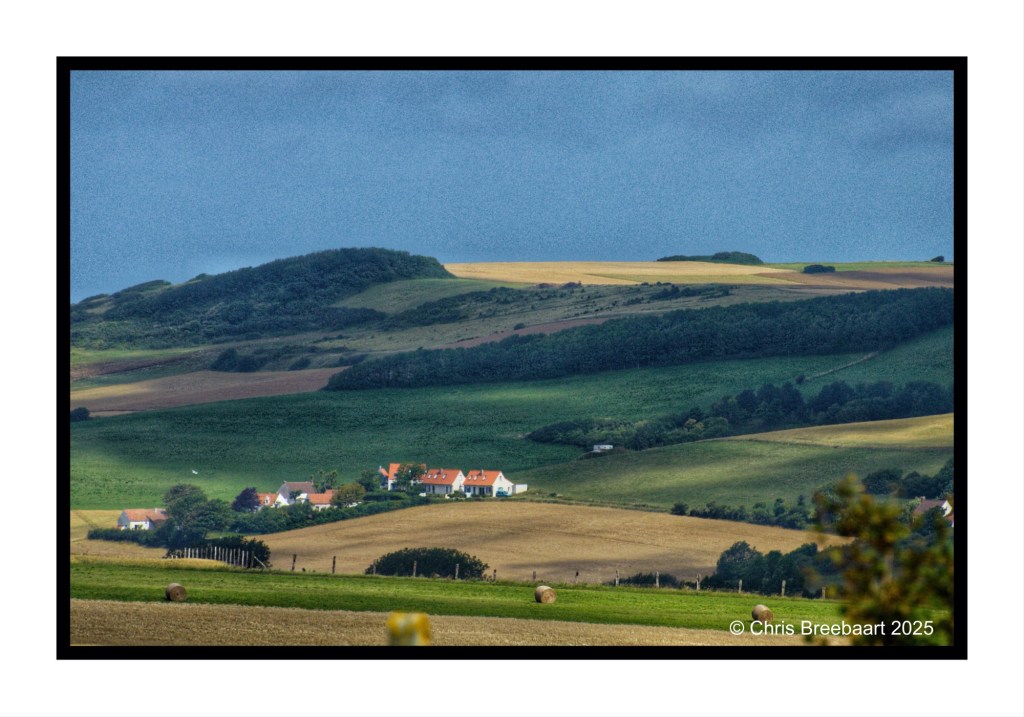 A scenic view of rolling hills and fields, with a small cluster of houses visible in the foreground, under a cloudy sky.