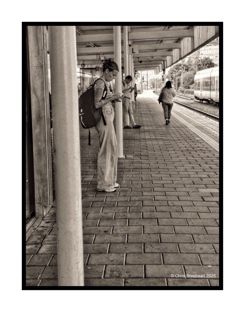 A group of people waiting at a train platform, some using their mobile phones, in black and white.