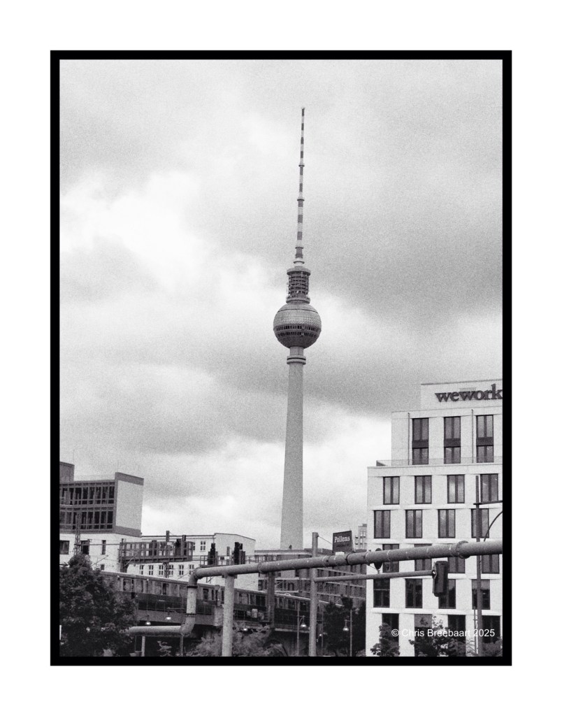 Black and white photo of the Berlin TV tower, known as Fernsehturm, surrounded by buildings and cloudy skies.