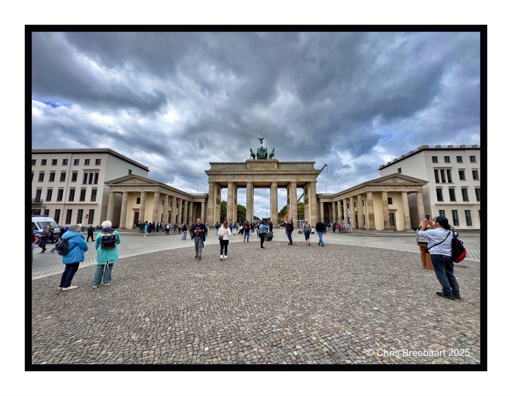 View of the Brandenburg Gate with pedestrians and cyclists in front, set against a cloudy sky.