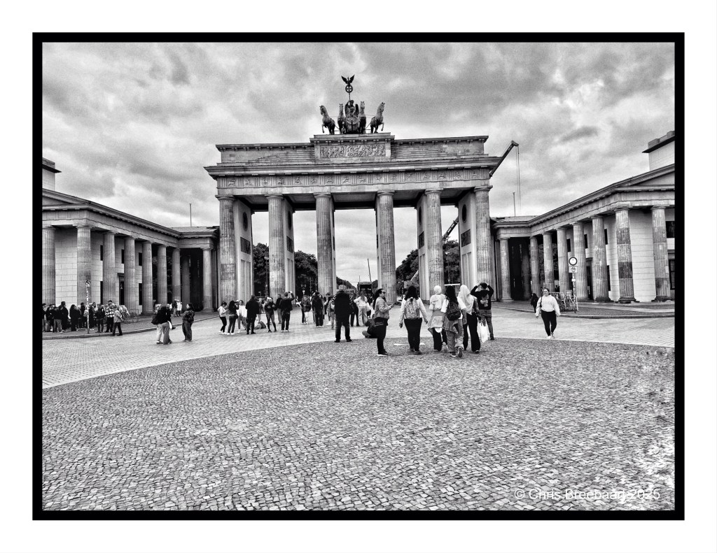 Black and white image of the Brandenburg Gate in Berlin, with a crowd of people gathered in front and historical architecture surrounding it.