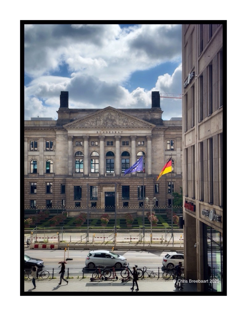 View of the Bundesrat building in Berlin, featuring flags and pedestrians in the foreground, with cloudy skies in the background.
