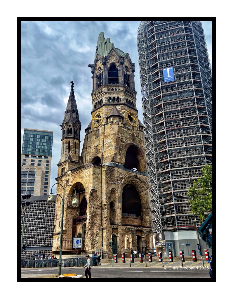 View of the Kaiser Wilhelm Gedächtnis Kirche, a historical church in Berlin, showcasing its damaged structure alongside modern buildings and scaffolding.