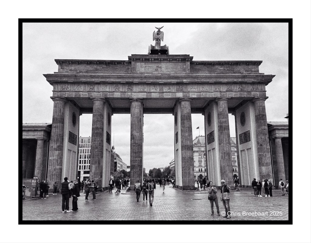 Black and white photograph of the Brandenburg Gate in Berlin, featuring a crowd of people walking through and around the iconic structure on a cloudy day.