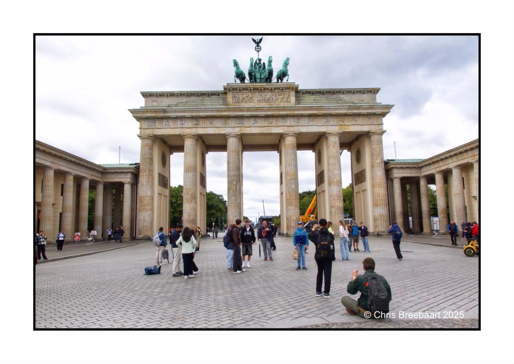 People gathered at the Brandenburg Gate in Berlin, showcasing the iconic monument surrounded by greenery and cloudy skies.