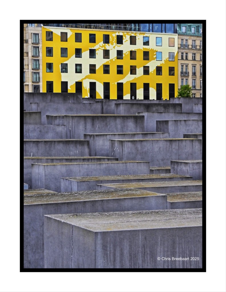 View of the Memorial to the murdered Jews in Europe, featuring abstract gray stone blocks in the foreground and a yellow building with artistic patterns in the background.