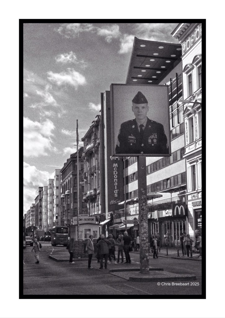 A black and white photograph of Checkpoint Charlie in Berlin, featuring a large portrait of a soldier on a billboard. The scene includes people walking and a McDonald's in the background.