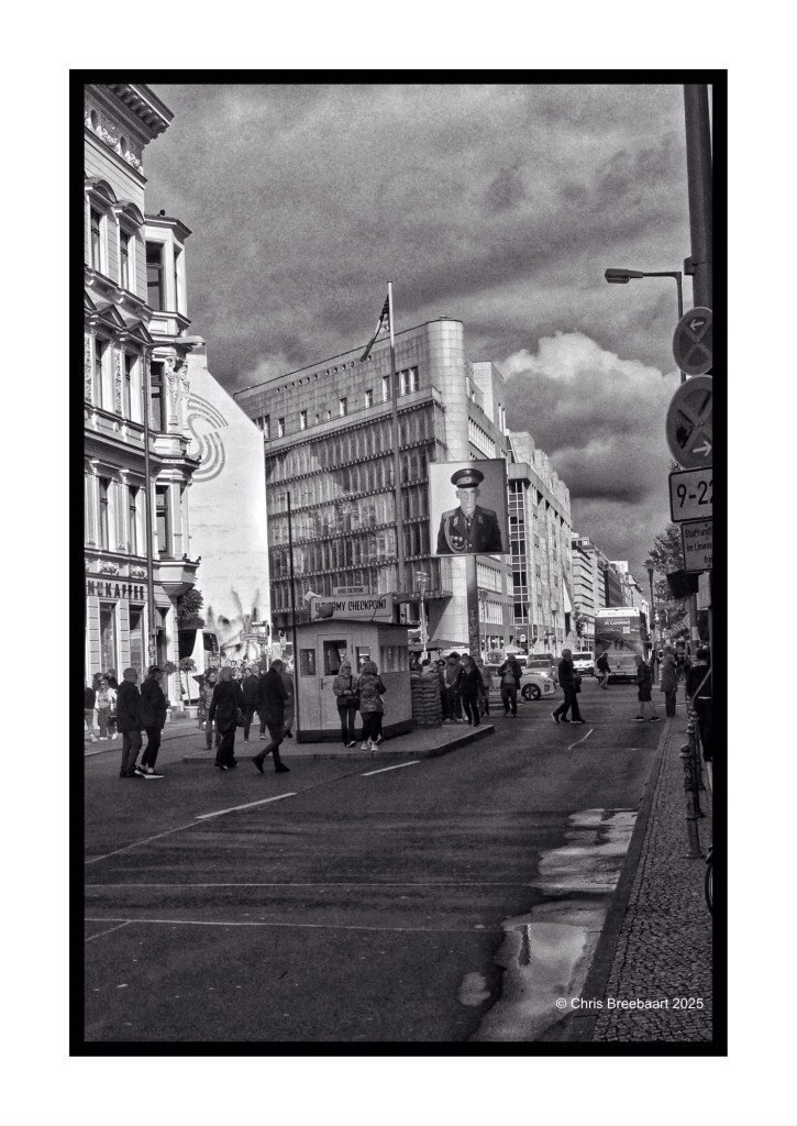 Black and white photograph of Checkpoint Charlie in Berlin, featuring a crowd of people walking past a historic guard booth and a large image of a soldier on a building.