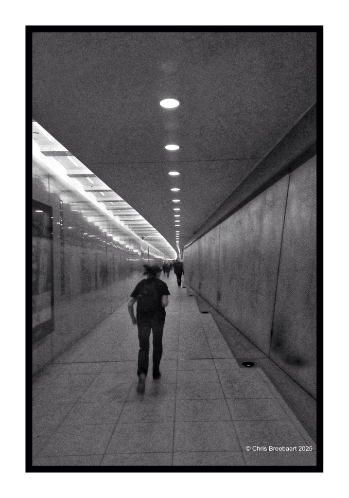 A black and white photograph of a subway corridor in Berlin, featuring a person walking ahead with another figure visible in the distance, illuminated by overhead lights.