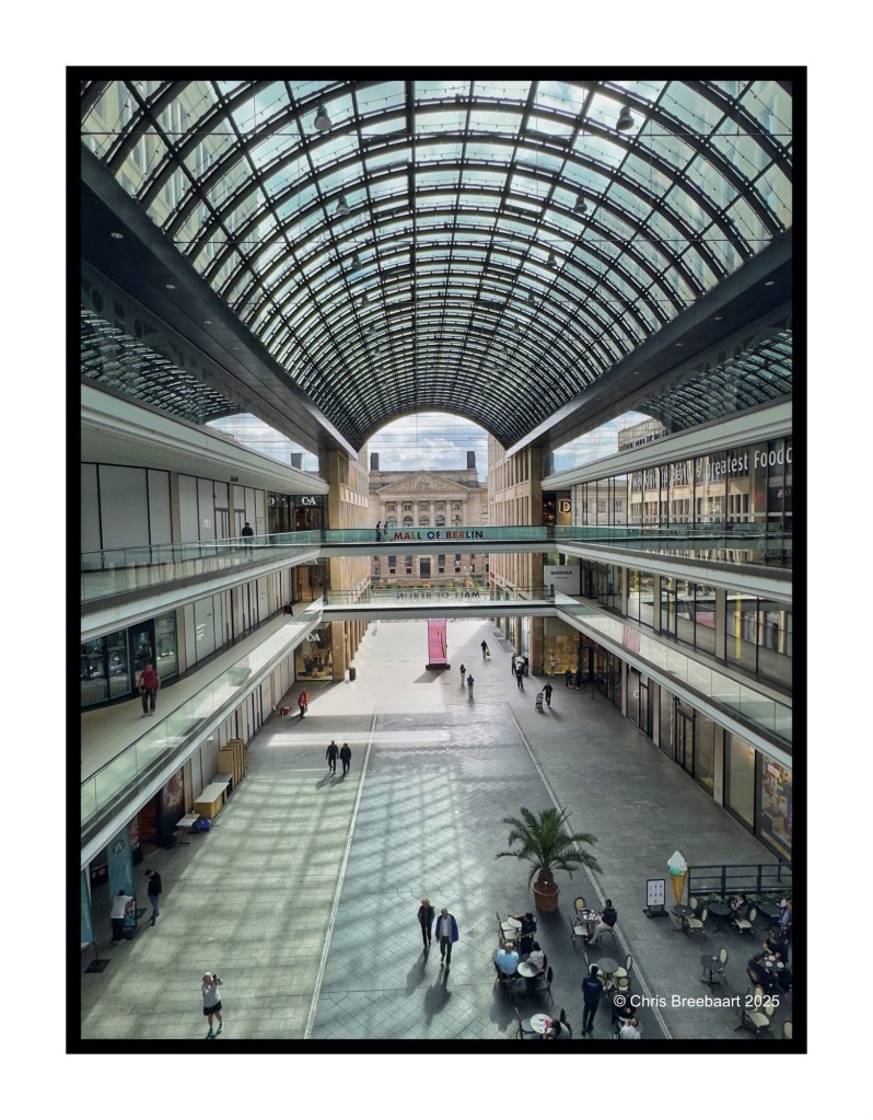 View of the interior of the Mall of Berlin, showcasing its glass roof and spacious design with people walking, shops lining the sides, and a food court area.