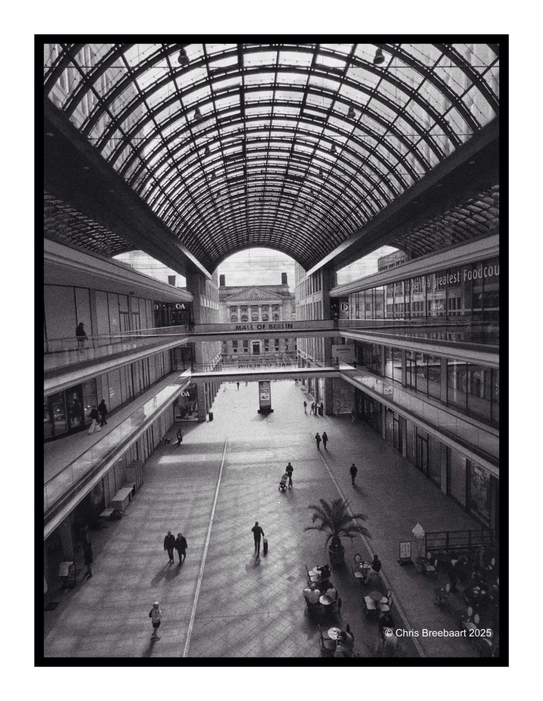 Black and white image showcasing the interior of the Mall of Berlin, featuring a high glass roof, shops lining the walkways, and people walking through the spacious atrium.