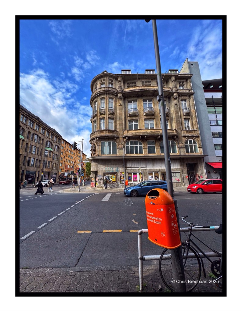 View of a historic building at the former Checkpoint Charlie in Berlin, with a blue sky and street scene, featuring a bicycle and an orange trash can.