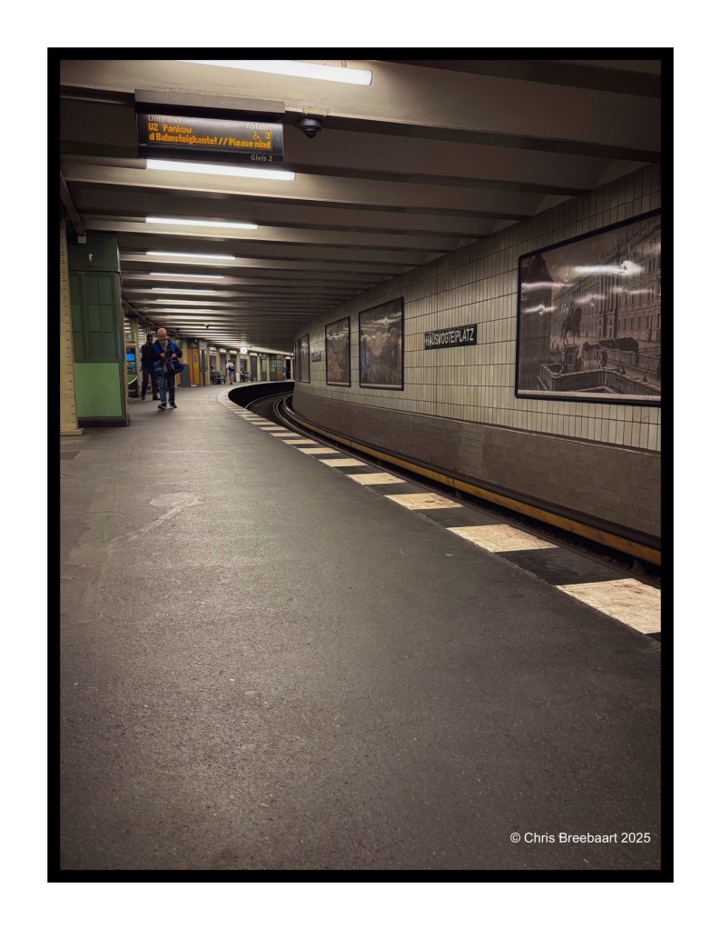 Interior view of the Hausvogteiplatz U-Bahn station in Berlin, featuring tiled walls, a curved platform, and train schedule display.