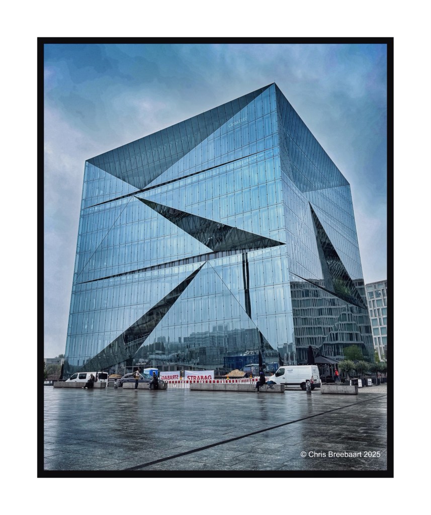 A modern glass cube building with angular reflective surfaces, set in an urban environment with vehicles and people in view. The sky is overcast, adding a dramatic effect to the scene.