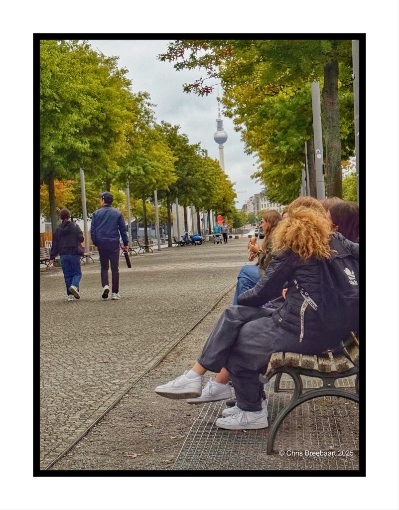 A view of the TV Tower (Fernsehturm) at Alexanderplatz, Berlin, with people sitting on a bench, surrounded by trees, and passerby walking in the background.