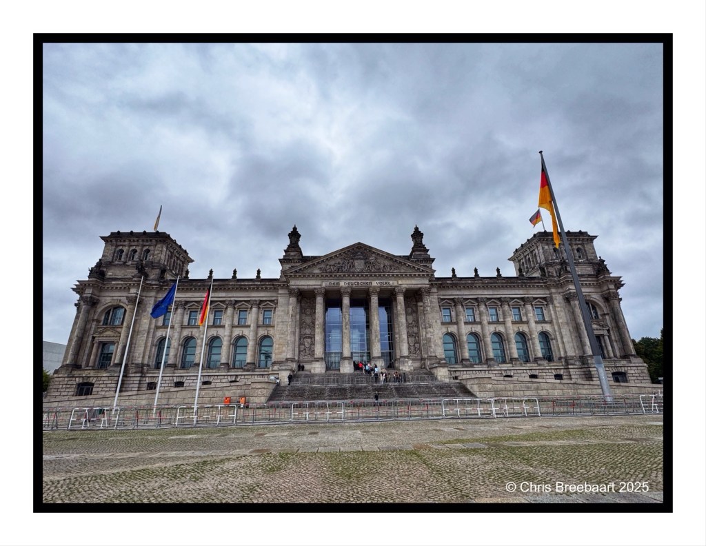 The front view of the Reichstag building in Berlin, showcasing its grand architecture and flags, with cloudy skies in the background.