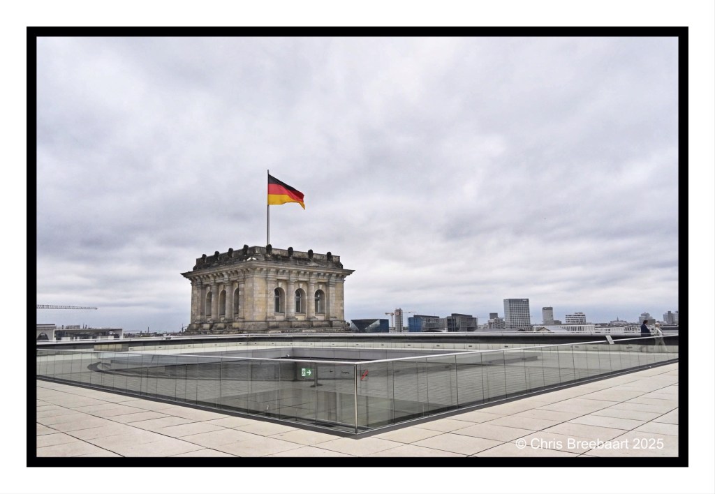 View from the roof of the Reichstag building in Berlin, featuring the German flag, a glass railing, and a cloudy sky.