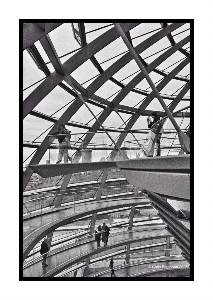A monochrome interior view of the Reichstag dome in Berlin, featuring several visitors walking along the curved glass and metal structure. The design showcases intricate geometric patterns, with reflections of the city visible through the transparent surfaces.