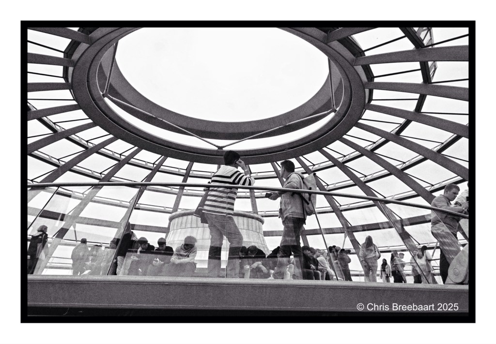Monochrome view from inside the Reichstag dome, showing visitors standing on a glass walkway surrounded by geometric architecture.