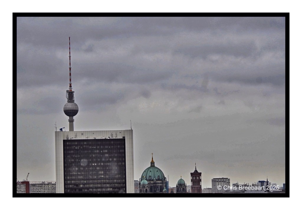 Panoramic view of Berlin's skyline, showcasing notable landmarks including the television tower, Berlin Cathedral, and the Red Town Hall against a cloudy sky.