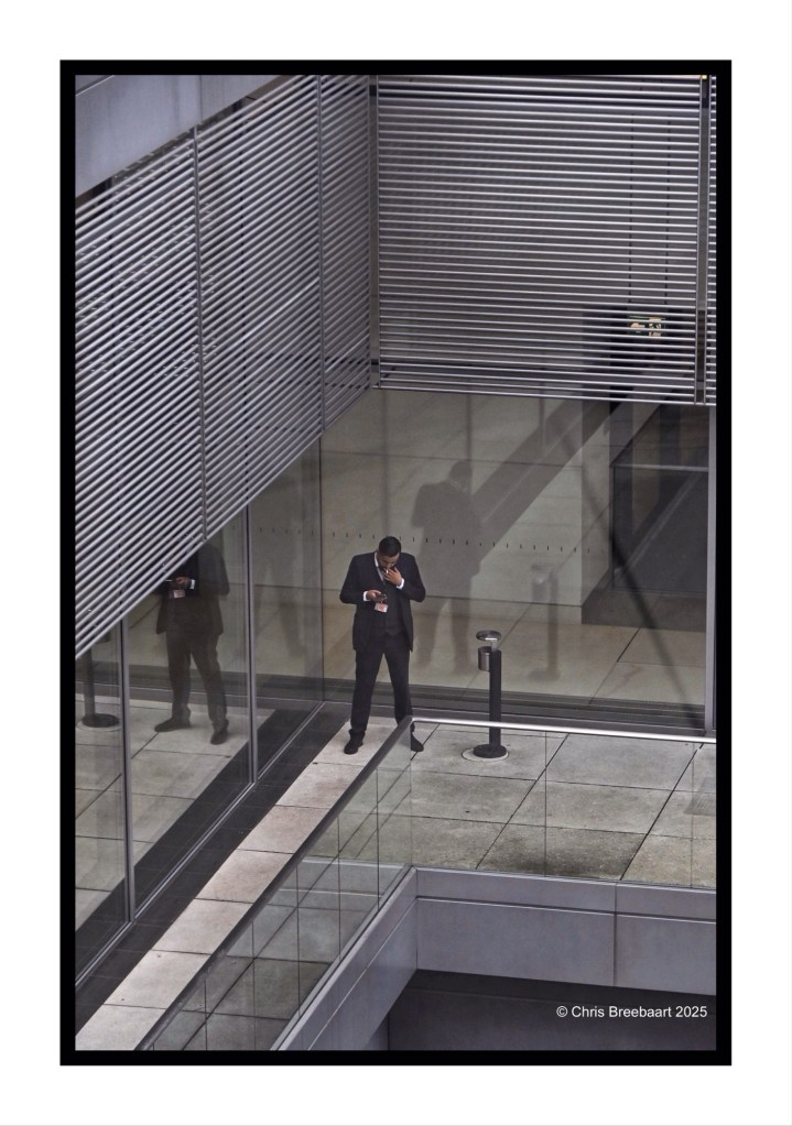 A staff member in formal attire standing on a terrace at the Reichstag, using a smartphone. Reflections of the building's modern architecture and venetian blinds are visible in the glass.