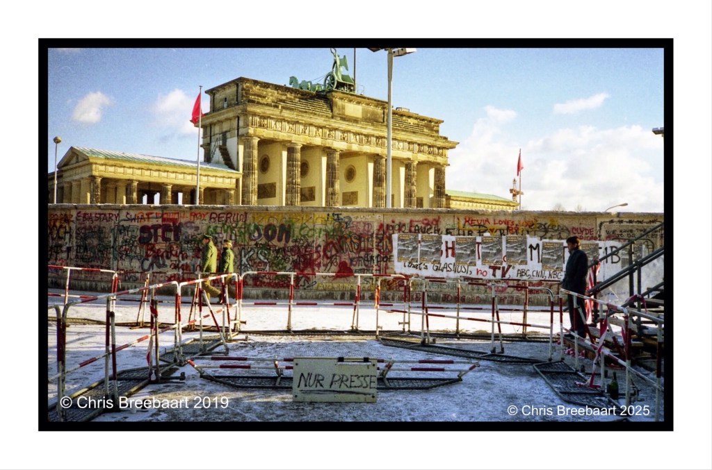 The Brandenburg Gate in Berlin, surrounded by a section of the Berlin Wall covered in graffiti, with people and barriers in the foreground.