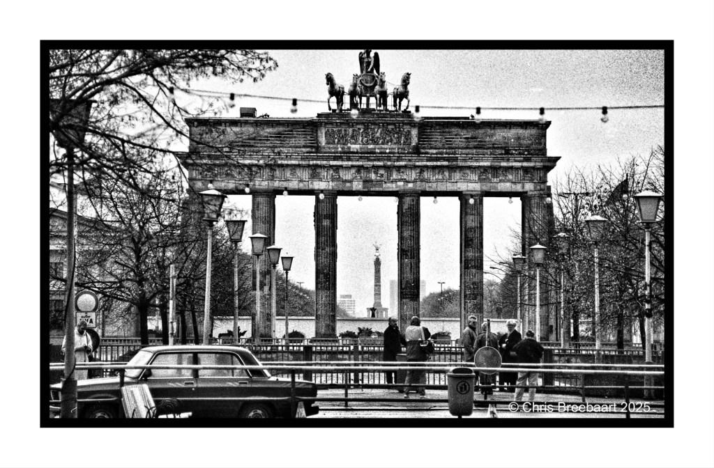 A black and white photo of the Brandenburg Gate in Berlin, framed by trees and streetlamps, with people walking beneath the iconic structure.