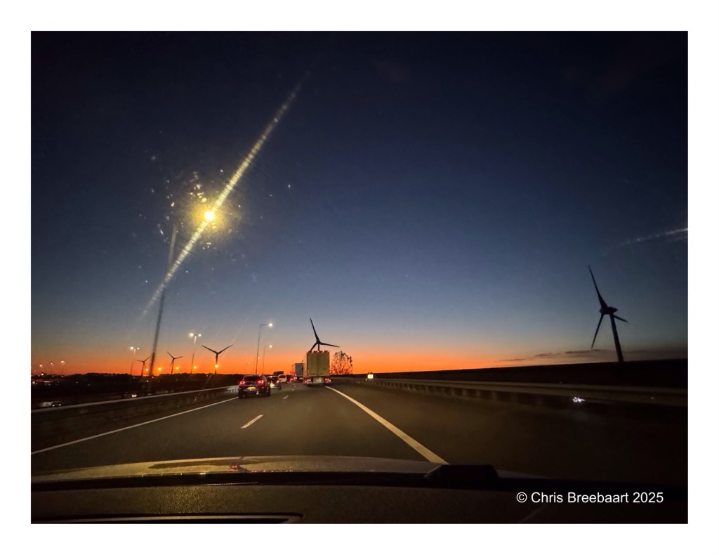 View of the A44 and A4 highways towards Schiphol Amsterdam at sunrise, with wind turbines along the roadside and a clear blue sky transitioning to orange hues.