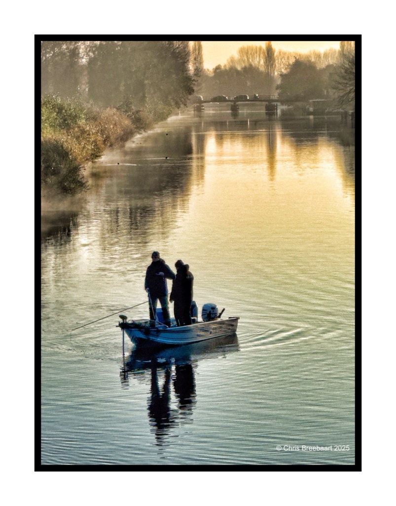 Three people fishing from a small boat on a calm river during an autumn sunrise, with reflections on the water and trees in the background.