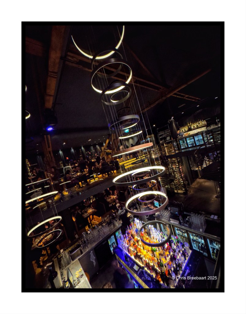 Interior view of the Encore venue in Scheveningen, featuring modern lighting fixtures and a vibrant bar area filled with colorful bottles. Diners can be seen enjoying their time in the stylish setting.