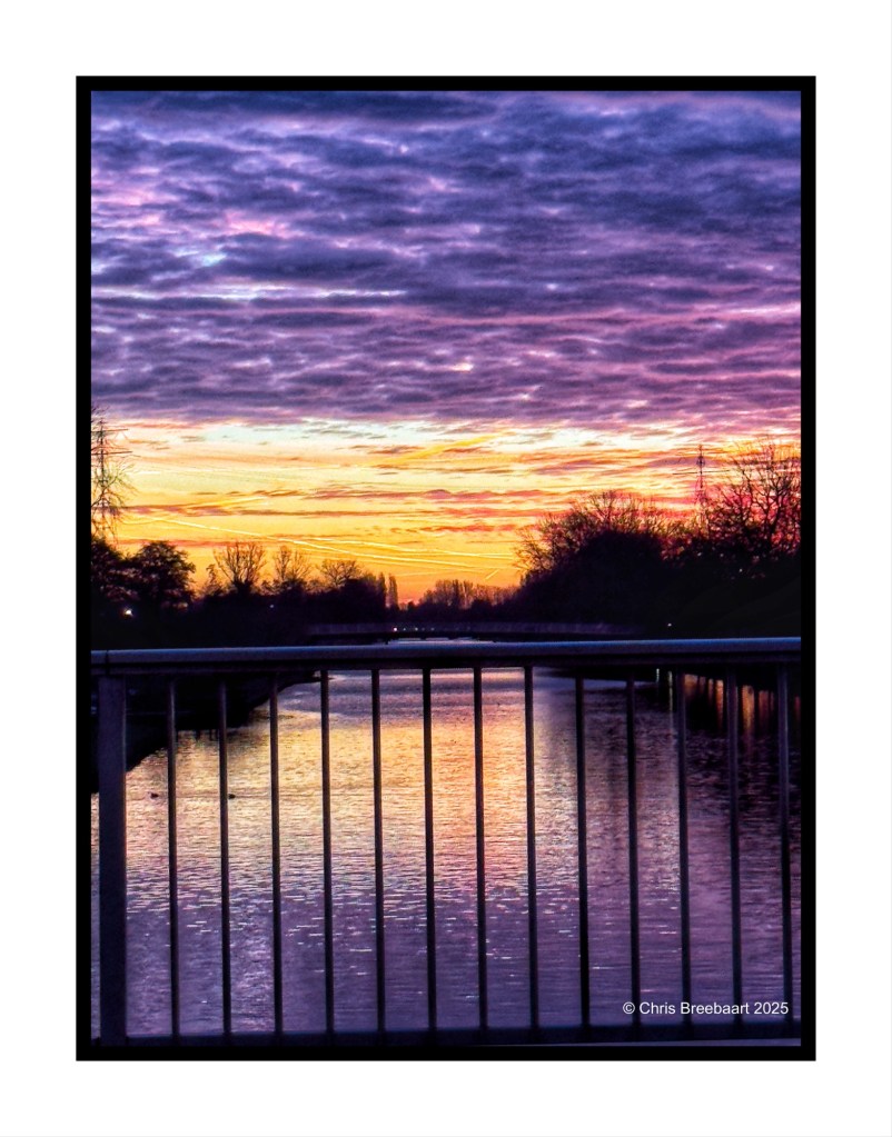 Autumn sunrise over a canal with colorful clouds reflected in the water, framed by a railing.