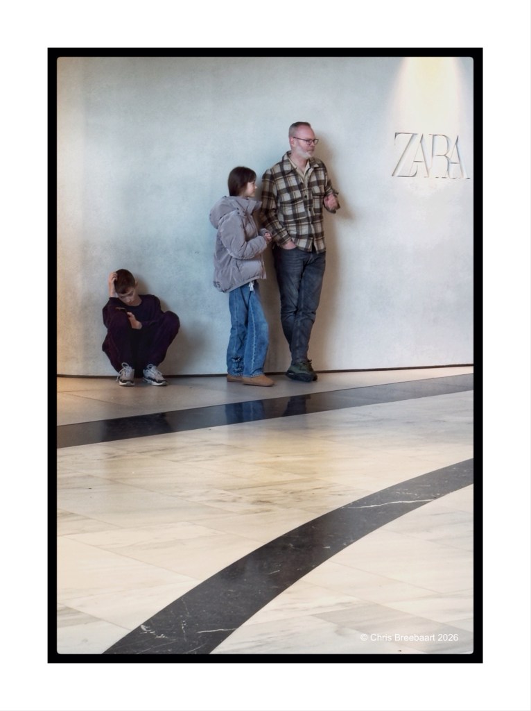A man and two children stand against a wall in a shop, with the Zara logo visible in the background. One child is sitting on the floor, while the other stands beside the man, both looking at him.