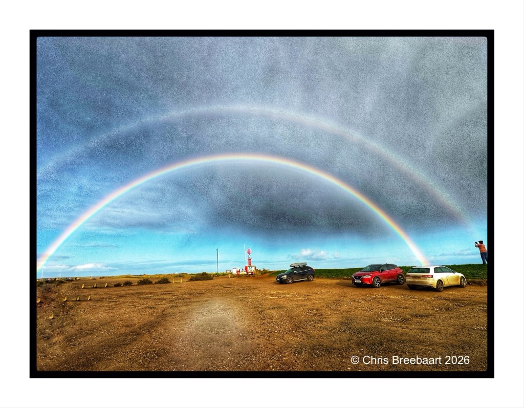 A scenic landscape featuring two rainbows in a cloudy sky, with several parked cars and a person photographing the scene in the foreground.