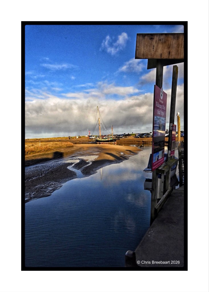 A serene coastal scene featuring a calm river reflecting the sky, with a sailboat in the background and a sandy shoreline. Signposts are visible on the right.
