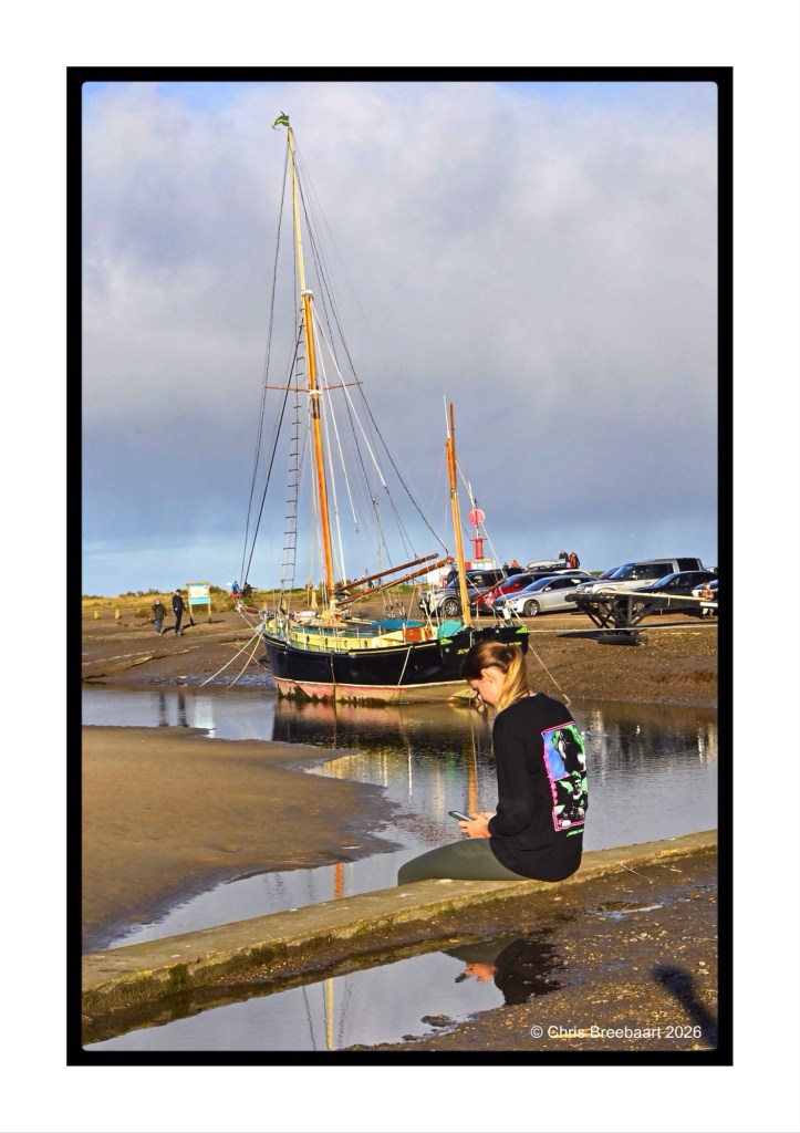A person sitting on a stone ledge near a calm body of water, looking at their phone, with a sailboat moored in the background and cars parked nearby.