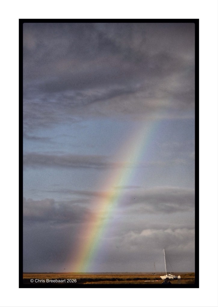 A rainbow arcs across a cloudy sky over a serene landscape with a small white sailboat in the foreground.