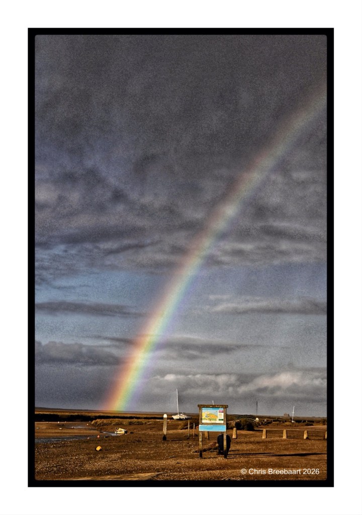 A vibrant rainbow arcs over a cloudy sky at a coastal location, with a signboard and boats visible in the foreground.