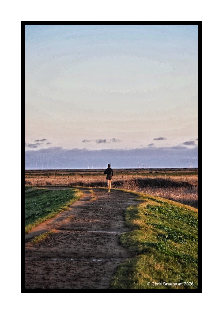 A person jogging on a dirt path leading towards a scenic landscape with grass and a distant view of wind turbines under a partly cloudy sky.