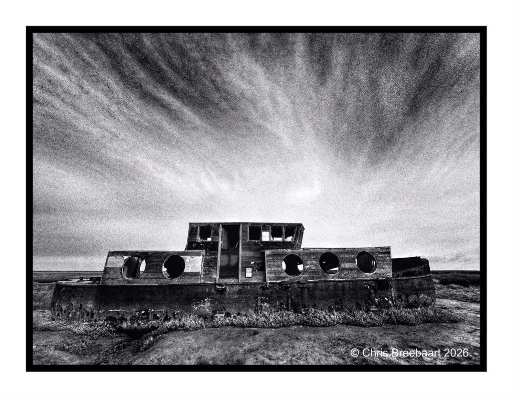 Black and white photograph of a dilapidated shipwreck on a barren landscape under a cloudy sky.