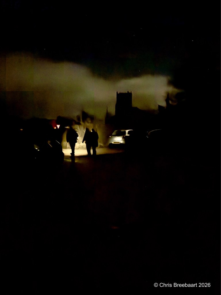 Two silhouetted figures walking on a dimly lit street at night, with a faint light illuminating their path, and a distant building silhouette against a darkened sky.