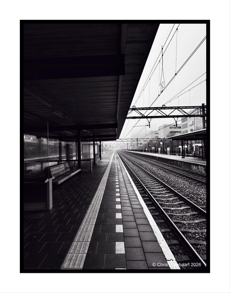 Black and white photograph of a deserted railway station platform with rail tracks extending into the distance, featuring a bench and clear sky.