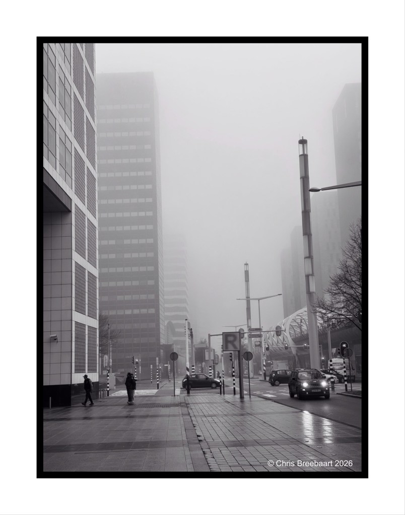 A foggy urban street scene featuring tall buildings, with pedestrians walking and cars on the road. The atmosphere is muted in black and white, enhancing the sense of depth and mystery.