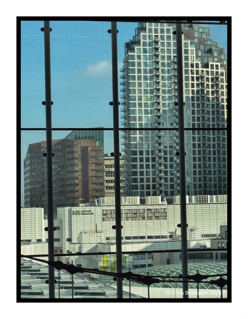 View of modern urban buildings through large glass windows, featuring a blend of different architectural styles and heights, with a clear blue sky in the background.