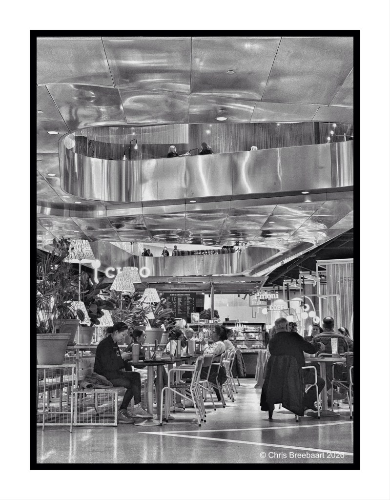 Interior view of a modern cafe with polished metal accents, featuring customers seated at tables and a balcony above with people. The space is styled with greenery and decorative lighting.